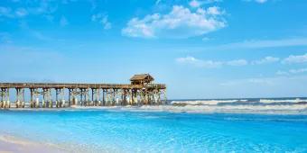 A long wooden pier with a shack at the end of it leading out to crystal clear water from a white sand beach.