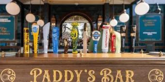 A wooden sign with Paddy's Bar engraved on it and two circular designs. Above there is a wide selection of different beers on tap.