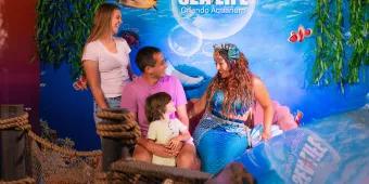 Mermaid with blue tail and seashells in her hair meeting a family including parents and a small kid. In the background there is a SEA LIFE sign.
