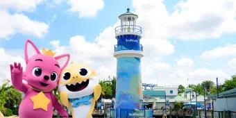 SeaWorld Summer Two mascot animals, one pink dog creature wearing a yellow crown. They are stood in front of a blue lighthouse on the rocks.