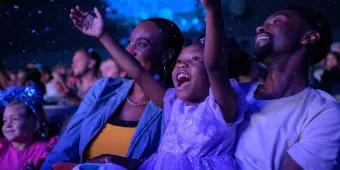Frozen Sing Along Celebration A little girl smiling and holding her arms up while sat with her mum and dad in a theatre
