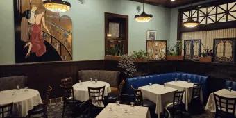 The Chef’s Table at The Edgewater Large blue room with dark decor, paintings and tables with white tablecloths inside.