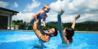A family enjoying a high-energy "darecation" moment in a sunny outdoor pool, with water splashing as a father lifts a young child
