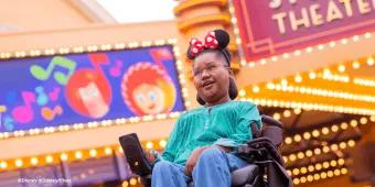 Guest in a wheelchair wearing Minnie Mouse ears smiling beneath the Studio Theater marquee at Production Courtyard, Disney Adventure World