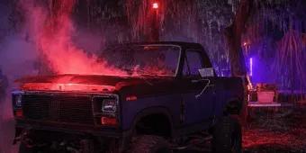 A dark pickup truck with red smoke rising from the open hood parked in swampy woods filled with neon lighting