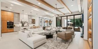 A spacious modern open-concept living room and kitchen featuring wooden ceiling beams, a large white sectional sofa, and floor-to-ceiling glass patio doors