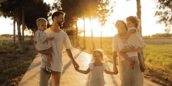 A happy family of five holding hands and carrying young children while walking along a paved path at sunset