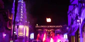 A crowd of guests walking toward the brightly lit Halloween Horror Nights entrance gate with bursts of fire in the night sky