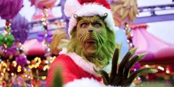 The Grinch, wearing a red Santa suit and hat, poses with a surprised expression in a brightly decorated theme park area