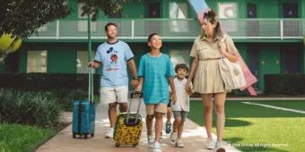 A family smiling and walking with their rolling luggage outside a green resort building