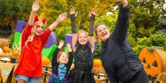A woman taking a selfie with three young girls dressed in Halloween costumes, with pumpkins and hay bales in the background