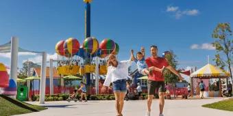 A mother and father swinging their young son by his arms while walking in front of a Peppa Pig balloon ride at a theme park