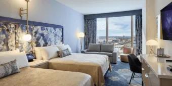 A standard hotel room featuring two queen beds with blue and purple floral patterned headboards, a sofa, and large windows with outdoor views