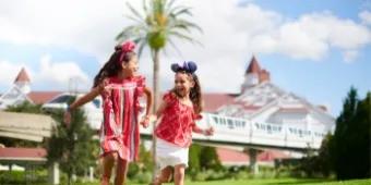 Two little girls wearing Mickey Ears running over grass while holding hands and laughing, with Disney's Grand Floridian in the background