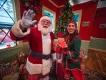 Christmas Town at Busch Gardens Santa waving at the camera standing next to one of his elves who is holding a present. They are standing in front of a Christmas tree