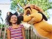 Timone with guest in Disney's Animal Kingdom Theme Park A girl smiling with eyes closed in front of the Tree of Life wearing glittery gold Mickey ears with a black bow on them. Stood next to her is Timone from the Lion King.