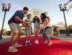 Family jumping on the red carpet in front of the large white stone entrance sign with Universal Studios Hollywood logo on. The family has dad on the right, one girl and one boy in the middle jumping and the mum crouched on the right.