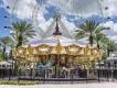 ICON Park A gold and white carousel with different pastel coloured horses and gold bars, in front of 6 palm trees diagonally lined behind and a huge ferris wheel which is white.