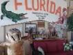 Florida huge welcome to florida sign on the wall with image of a gator on and below there is a statue of an alligator stood up smiling and waving. Next to the statue there is an old sofa with old images above.