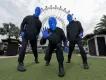 Blue Men Group at Icon Park There are three men completely painted in blue paint stood in front of a big white Ferris wheel on a field. Each of them are posing in a strange way.