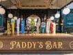 Paddy's Bar A wooden sign with Paddy's Bar engraved on it and two circular designs. Above there is a wide selection of different beers on tap.