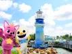 SeaWorld Summer Two mascot animals, one pink dog creature wearing a yellow crown. They are stood in front of a blue lighthouse on the rocks.