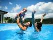 A family enjoying a high-energy "darecation" moment in a sunny outdoor pool, with water splashing as a father lifts a young child
