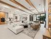 A spacious modern open-concept living room and kitchen featuring wooden ceiling beams, a large white sectional sofa, and floor-to-ceiling glass patio doors