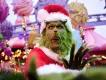 The Grinch, wearing a red Santa suit and hat, poses with a surprised expression in a brightly decorated theme park area