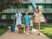 A family smiling and walking with their rolling luggage outside a green resort building