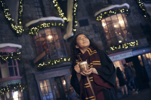 A young woman wearing a scarf and wizard robes drinking Butterbeer in front of decorated buildings