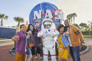A group of 5 friends posing with someone in an space suit in front of the NASA logo