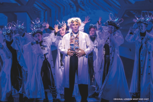 Doc Brown wearing a white lab coat standing centre stage in front of dancers also wearing lab coats