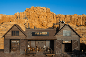 A wooden barn-like building in front of sandy cliffs with a carved Aztec face in the centre