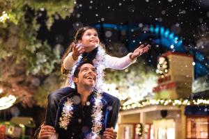 A girl sitting on a man's shoulders holding her hands out to catch fake snow as it falls