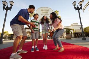Family jumping on the red carpet in front of the large white stone entrance sign with Universal Studios Hollywood logo on. The family has dad on the right, one girl and one boy in the middle jumping and the mum crouched on the right.