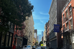 View down street with buildings on either side and cars. There is a person dressed in red walking along pavement. 