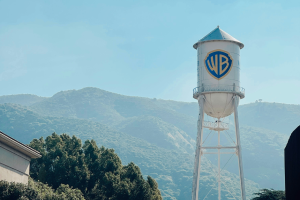 White water tower with blue shield shaped logo with letters WB on. In the background there is a large tree and big mountains with a blue sky overhead.