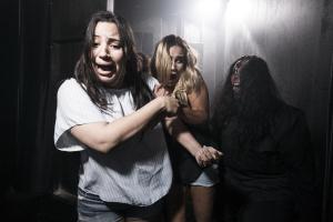 Two young women in a dark room with a bright light shining behind with terrified facial expressions, clutching onto one another and dodging another person who is lurking in the dark. The third person has black hair and a wound on their head which seems to be bleeding down their face. Their hair is messy and they are pulling a scary face with their tongue poking out.