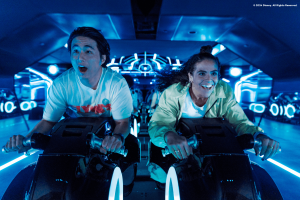 Two young adults sat on motorcycles shaped vehicles TRON Lightcycle. They are leaning forward gasping with shock. 