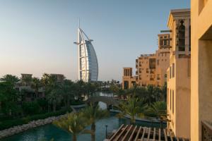 Beige buildings with a river beside them and some trees. It is sunset and the view shows the Burj Al Arab, a ship sail shaped building.