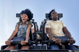 Two people screaming on a rollercoaster with only lap bars holding them in. They are looking down in shock ready to fall.
