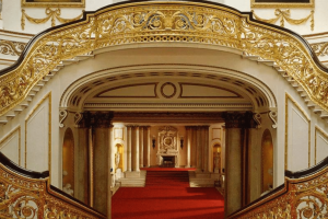 Golden and wooden bannisters lining two staircases which wrap around and join to create on large staircase. There is bright red carpet and white walls lined with gold details. There is a large open hallway with a fireplace at the end.