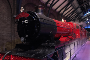 Large steam train with the label Hogwarts Express, coloured black and a dark red. There is steam surrounding and it is parked in a large factory type building.