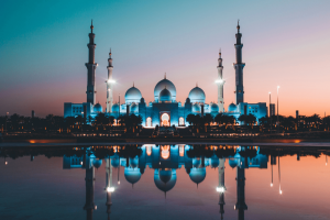 A beautiful religious temple in Abu Dhabi located overlooking a lake. The sun is setting with a pink and blue hue in the sky and the temple is perfectly reflected in the lake. It is a white building with three white domes and four big pillars sticking out.