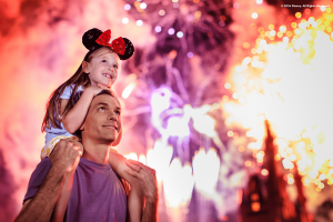 Young girl sat on her dads shoulders as the both look into the sky watching the magnificent fireworks above Cinderella Castle. There are many different colours and lights.