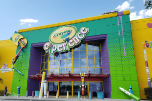 Large modern building coloured yellow, green and purple has big sign saying Crayola Experience on it. There are giant crayons outside with faces on them.