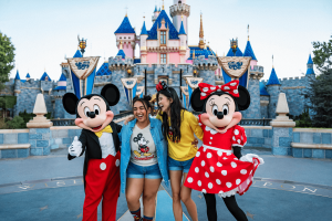 Two young women stood with Mickey and Minnie in front of a pink and blue castle.