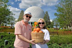 Man and woman holding a basket full of oranges in front of the giant golf ball looking structure and green grass and trees.