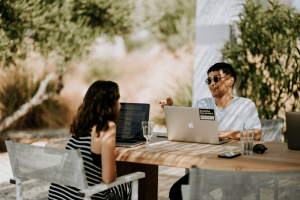 A woman and man sat at a wooden table outside talking in the sun with their work laptops open.